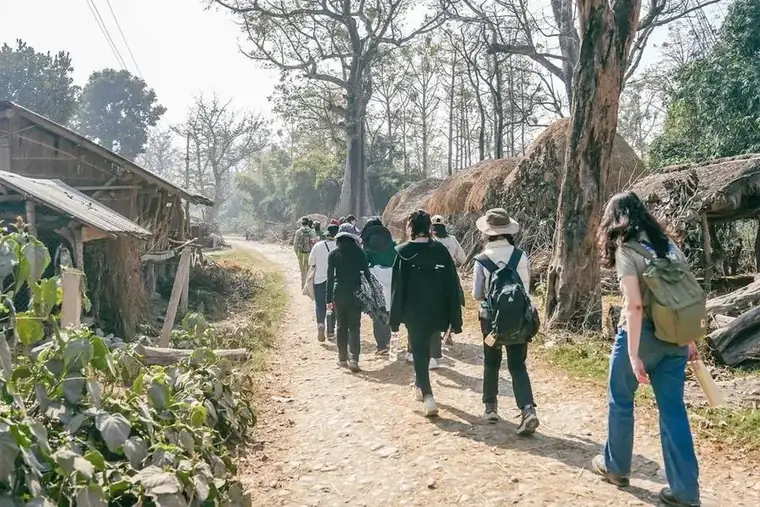 Students walking a track into a Tharu village past thatched longhouses