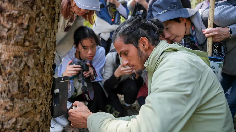 A Nepali field guide fitting a camera trap to a tree while students crouch round it with cameras