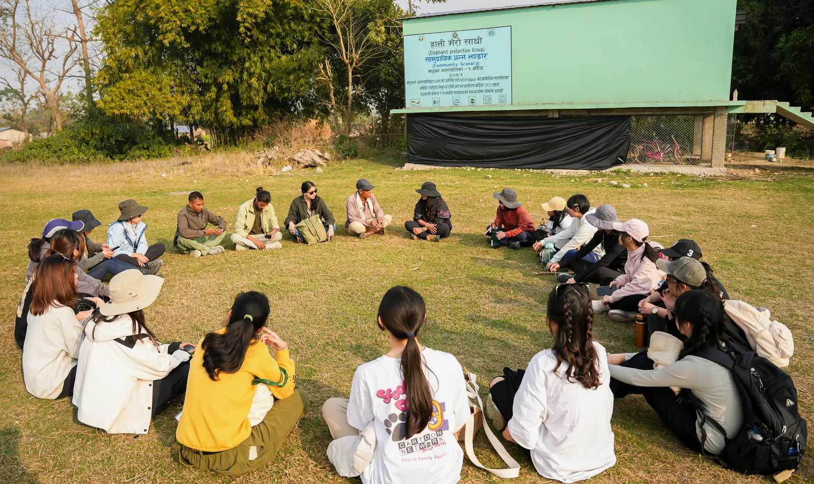 Students and community members sitting in a circle on the grass in front of the Elephant Protection Group community granary and its projection screen in Bardiya